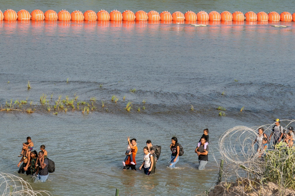 مهاجرون يسيرون بين الأسلاك الشائكة وسلسلة من العوامات في نهر ريو غراندي في إيجل باس، تكساس (أ ف ب)   